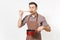 Young smiling man chef or waiter in striped brown apron, shirt tasting red empty stewpan, wooden spoon isolated on white