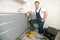 Young smiling man plumber in uniform fixing the sink with adjustable spanner in his hand working in the kitchen