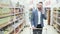 Young smiling man in white shirt is talking on smartphone and walks between shelves with goods in supermarket, steadicam