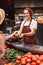 Young smiling seller in apron behind counter with vegetables while happily giving bananas to customer in modern