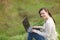 Young smiling successful smart business woman or student in light casual clothes sitting on grass using laptop in field