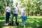 young smiling volunteers with new trees rake watering can and shovel standing in green park