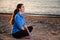 A young smiling woman in sportswear is sitting cross-legged on the sand. In the background the sea. Side view. Wellness