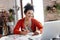 Young smilng woman with dark curly hair sitting at the table happily working on laptop drawing fashion illustrations