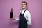 Young sommelier guy holds a bottle of red wine on a colored background, an expert on wine in a white shirt and apron, the waiter