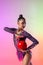 Young sportive girl, rhythmic gymnast posing with red ball isolated on colored neon background