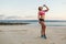 young sportswoman in earphones with smartphone in armband case drinking water from bottle on beach with sea