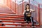 Young sporty beautiful woman having a break sitting on the red stairs after the morning jogging in the modern city, checking