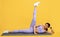Young sporty black lady warming up and lifting leg up, exercising on mat over yellow background