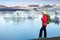 Young sporty girl with backpack admires beautiful view of the ice lake and mountains. Ice lagoon in Iceland