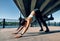 Young sporty woman doing yoga asana downward facing dog under industrial bridge