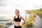 Young sporty woman runner in black activewear standing outside by the seaside, resting.