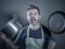 Young stressed and funny lazy man with apron holding kitchen pan and kitchen pot screaming in stress desperate crying isolated bac