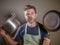 Young stressed and funny lazy man with apron holding kitchen pan and kitchen pot in stress desperate crying isolated background