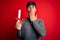 Young student man holding university graduated diploma degree over red background cover mouth with hand shocked with shame for