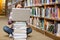 Young student sitting on library floor using laptop on pile of books