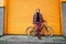 Young stylish hipster man standing with a cruiser city Bicycle near orange Wall. Student Resting after ride bicycle. Eco