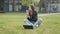 Young stylishly dressed girl sitting in the park on the grass near the computer, holding a dandelion and blowing flowers