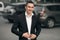 Young successful smiling man in a black suit, on the street, on the background of cars. Happy modern man. Outdoors