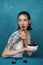 Young surprised woman sits at the table with cherry bowl in front of blue background