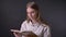 Young sweet girl is reading book, watching at camera, grey background