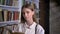 Young sweet girl is reading book, watching at camera, library on background