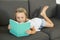 Young sweet and happy little girl 6 or 7 years old lying on home living room sofa couch reading a book quiet and adorable in child