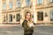 Young talented female student with broad smile in casual clothes standing in front of university building holding books and coffee