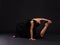 A young and talented girl gymnast, sits in one knee and bends back. On a black background.