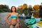 A young tanned woman in a bikini poses on the river bank. In the background, a man sitting on a kayak. Active recreation