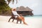 Young tanned woman sit at the chair and relax near bungalow of resort on the beach. Happy woman vacation. Island lifestyle. Rest