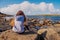 Young teenager girl on rough stone coast looking at the ocean, back to viewer. Life in tough conditions concept. Warm sunny day,