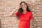 Young teenager girl standing over bricks wall doing peace symbol with fingers over face, smiling cheerful showing victory