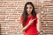 Young teenager girl standing over bricks wall in hurry pointing to watch time, impatience, looking at the camera with relaxed