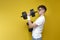 Young thin, not athletic, nerd guy lifts heavy dumbbells and shows strength on a yellow background