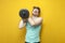 Young tired thin guy nerd lifts heavy dumbbells and cries on yellow background, confused student in glasses is training