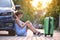 Young tired woman with suitcase sitting near her car waiting for someone. Travel and vacations concept