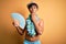 Young tourist man on vacation wearing swimwear and hawaiian lei flowers holding hand fan cover mouth with hand shocked with shame