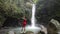 A young tourist standing at a waterfall, capturing photos using a smartphone.