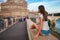 Young tourist woman sitting on the ancient embankment Tiber in Roma at sunset. Near of bridge and castle of Angels. Saint Angelo