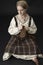 A young Viking woman wearing a plaid tunic sits on the ground sewing a leather bag