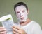 Young weird and funny man at home trying using beauty paper facial mask cleansing learning anti aging facial treatment reading the