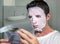 Young weird and funny man at home trying using beauty paper facial mask cleansing learning anti aging facial treatment reading the