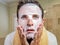 Young weird and funny man at home trying using beauty paper facial mask cleansing learning anti aging treatment in surprised  face