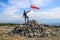 Young white Caucasian male tourist in sportswear proudly stands on a mountainous stone peak and holds a flag