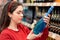 A young white woman examines a bottle of blue wine. In the background, store shelves are blurred. The concept of buying alcohol in
