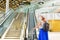 Young woman 40s in protective face mask with a suitcase riding escalator in airport terminal. Safety travelling alone