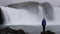 A young woman admires a powerful raging waterfall that falls heavily along a rocky edge. A crystal clear stream of glacier water f