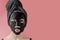 Young woman apply black cosmetic fabric facial mask on pink background. Face peeling mask with charcoal, spa beauty treatment,