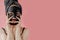 Young woman apply black cosmetic fabric facial mask on pink background. Face peeling mask with charcoal, spa beauty treatment,